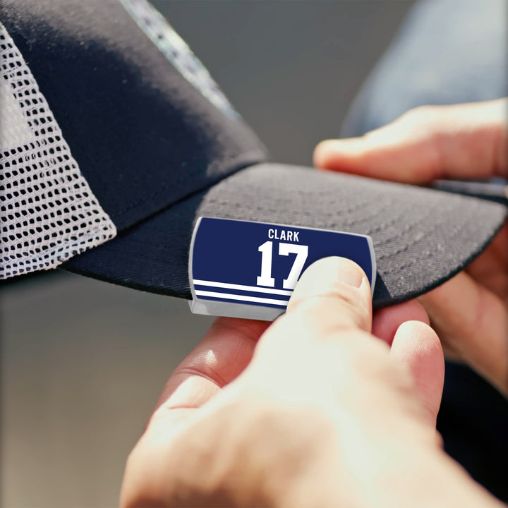 A close-up of hands attaching a ZippyBack™ Wendel Clark name tag with white stripes to the brim of a black and white mesh baseball cap, inspired by hockey legend Wendel Clark’s legacy.