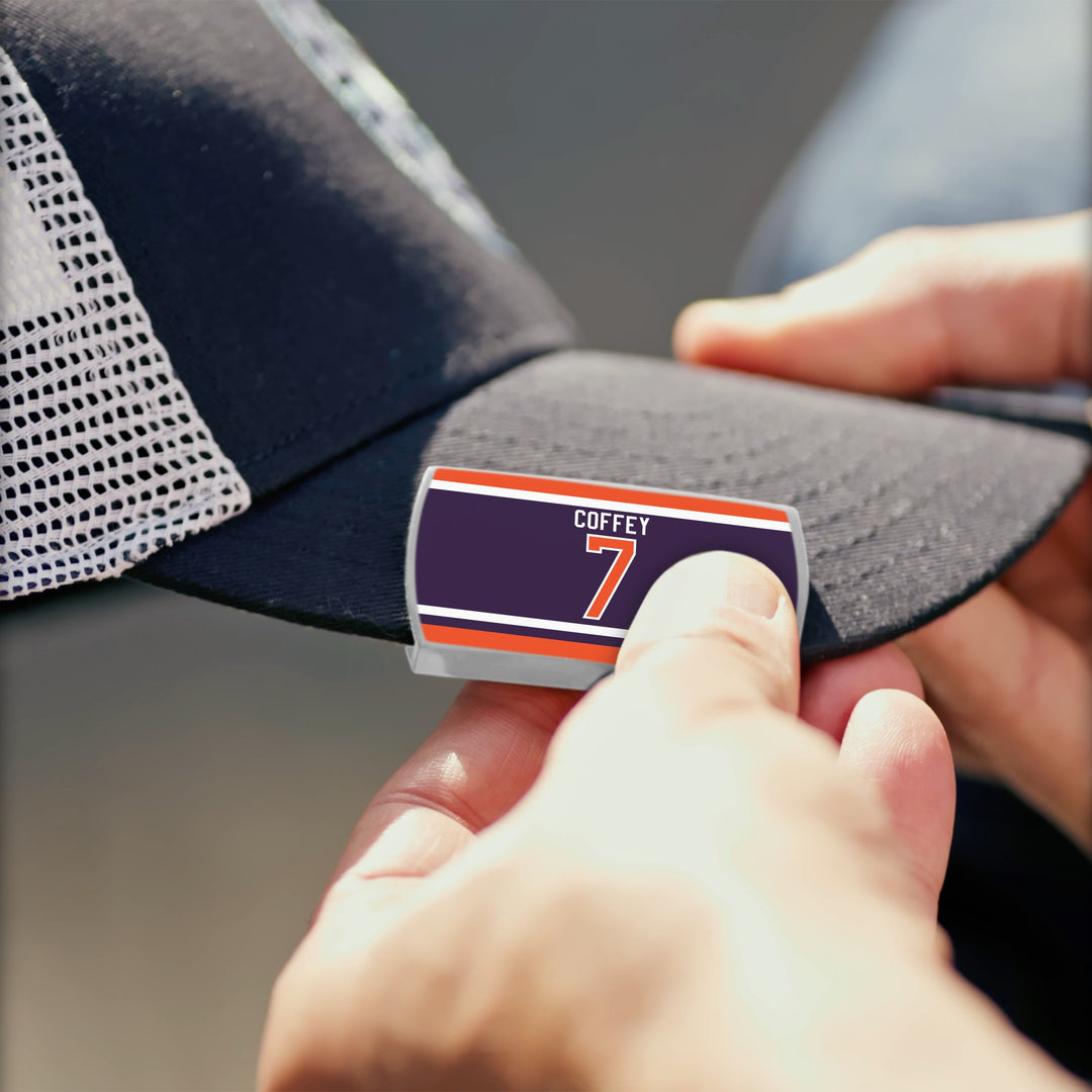 Close-up of hands placing a Limited Edition COFFEY 7 sticker with bold stripes on the brim of a black and white ZippyBack™ Paul Coffey mesh baseball cap from the Rafters Series 1.