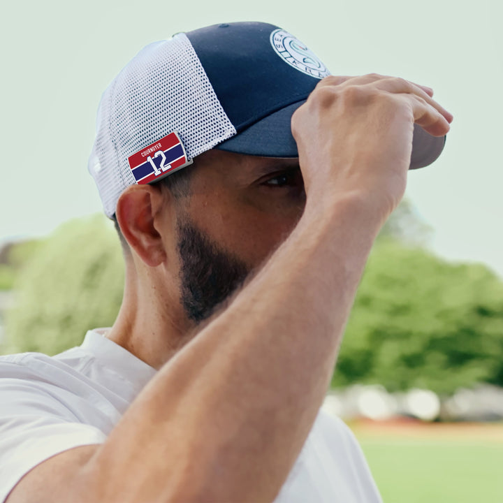 A man in a white shirt adjusts his ZippyBack™ Yvan Cournoyer mesh cap—navy blue and white with a small red "12" patch, honoring the hockey legend—while standing outdoors amid blurred greenery.