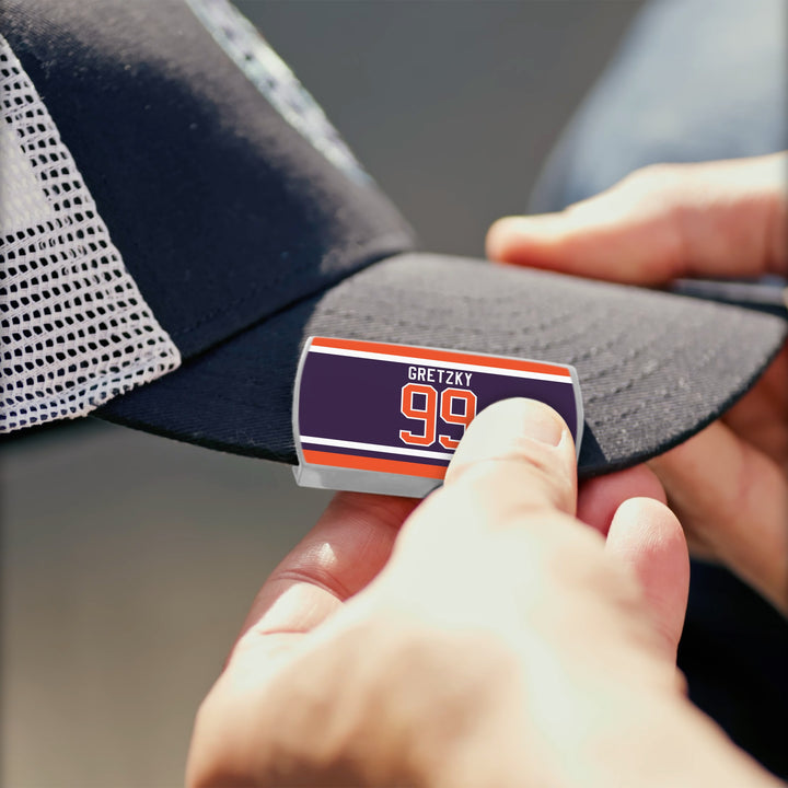 A person attaches a ZippyBack™ Wayne Gretzky Limited Edition sticker reading “GRETZKY 99” with orange and white stripes to the brim of a navy blue mesh cap, celebrating the hockey legend’s iconic number.