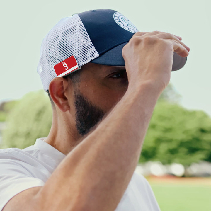 A bearded man adjusts the brim of a navy and white ZippyBack™ Gordie Howe mesh cap from Rafters Series 1. A red tag with a white g marks this Limited Edition cap. Blurred trees and grass are in the background.