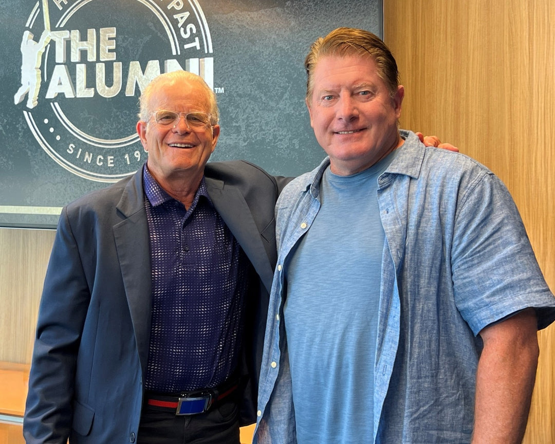 Two men stand smiling together in front of a sign that reads “THE ALUMNI.” One man wears a suit and glasses, while the other wears a casual blue shirt. They appear to be indoors in a well-lit room.