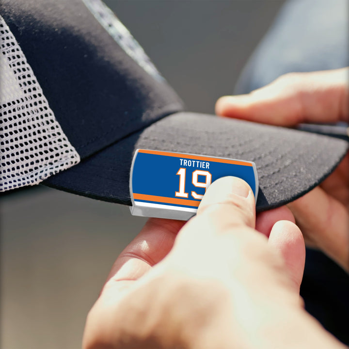 A person attaches a blue, orange, and white label with "Bryan Trottier" and the number 19 to the brim of a black and white ZippyBack™ mesh cap.