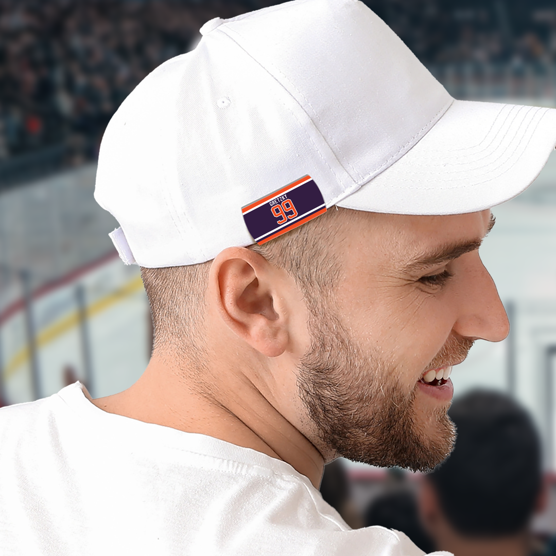 A man wearing a white cap and shirt smiles while attending a hockey game. The cap features a small rectangular patch with the number 99 in red on a blue background with orange and white stripes.
