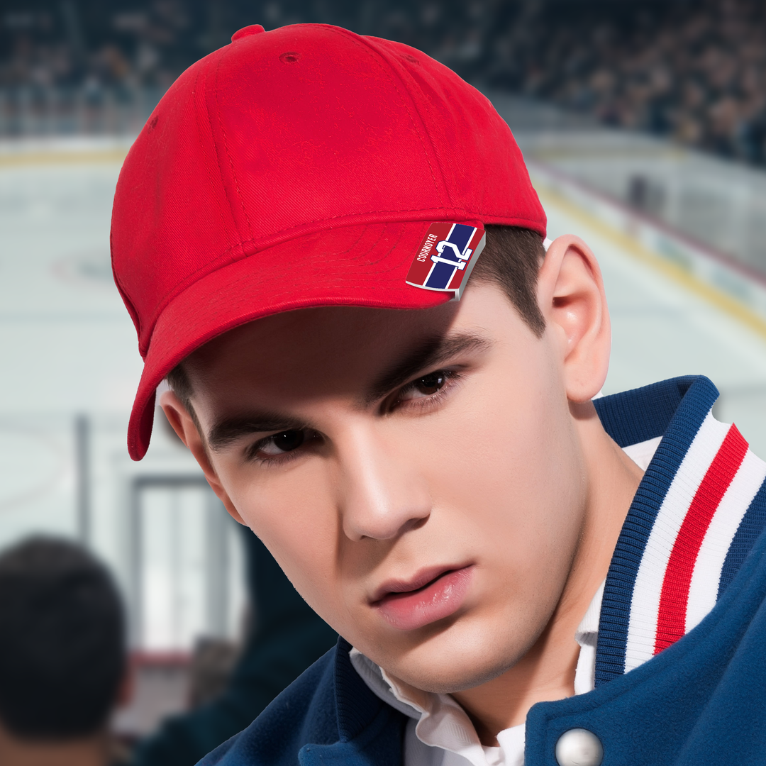 A young man wearing a red baseball cap and a varsity jacket sits in the stands at an ice hockey rink, with the rink and audience blurred in the background.