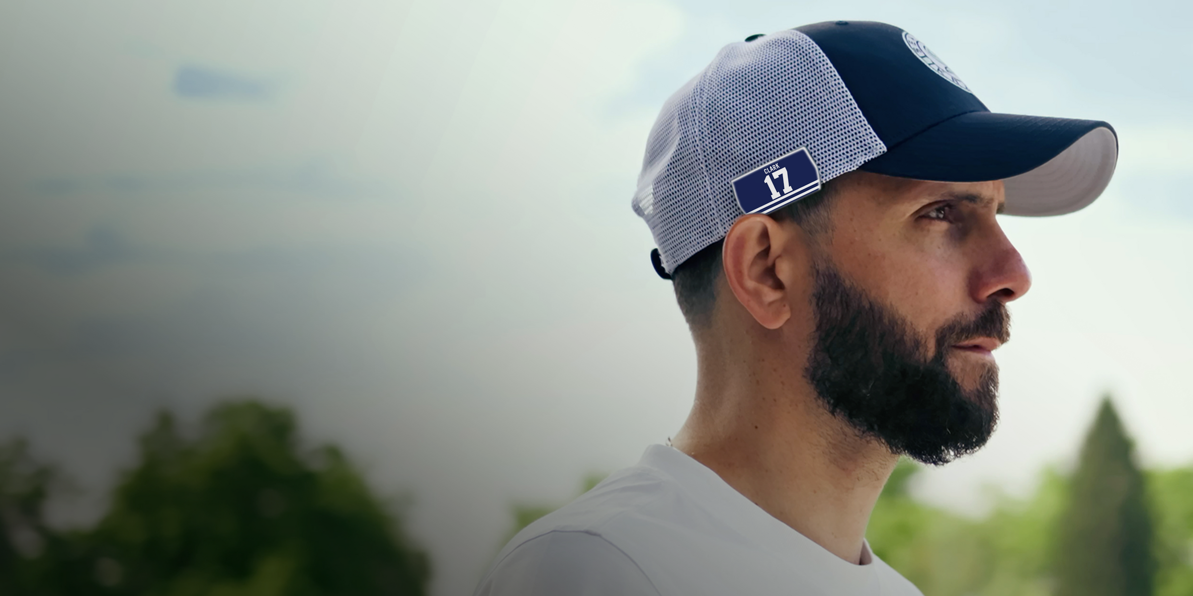 A man with a beard wearing a blue and white mesh baseball cap and a white shirt stands outdoors, looking thoughtfully into the distance with a blurred background of trees and sky.