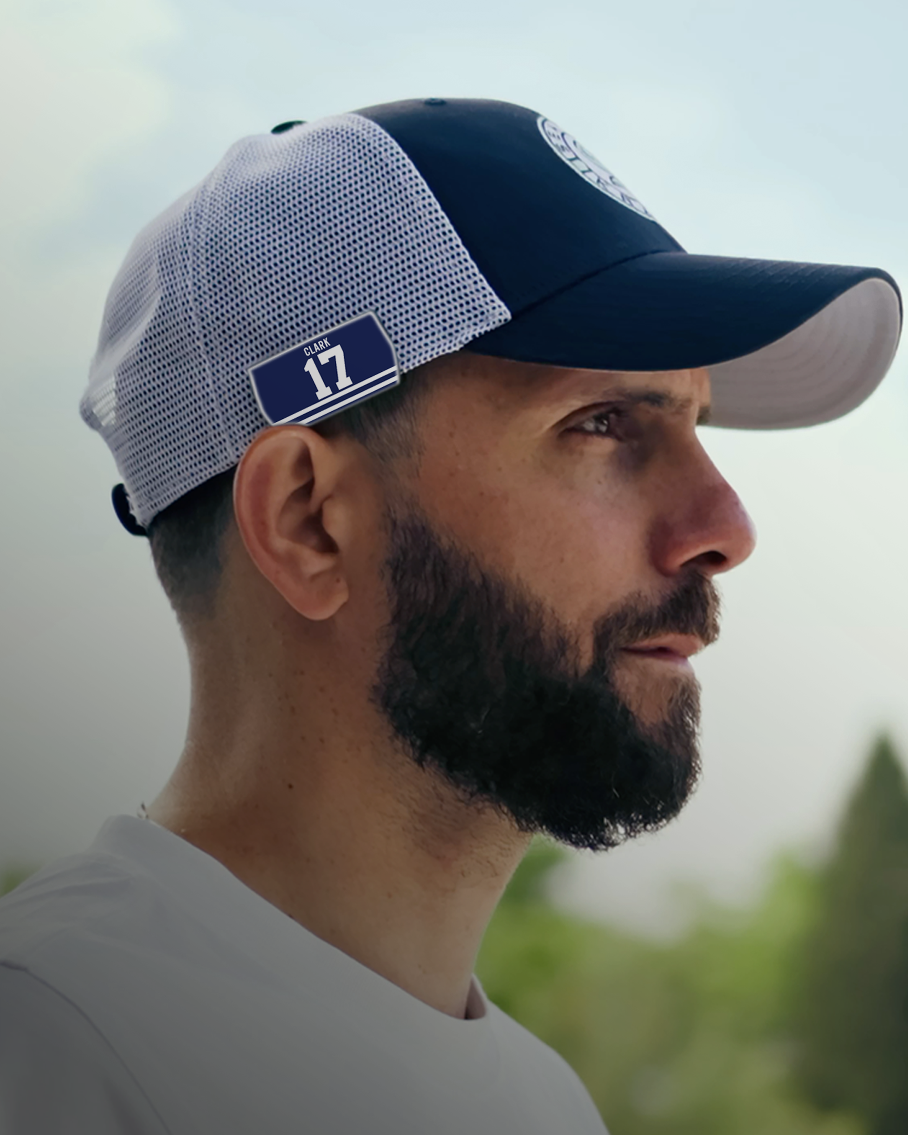 A man with a dark beard wearing a blue and white mesh cap with the number 17 on the side, looking to the right. The background is blurred with greenery and a light sky.