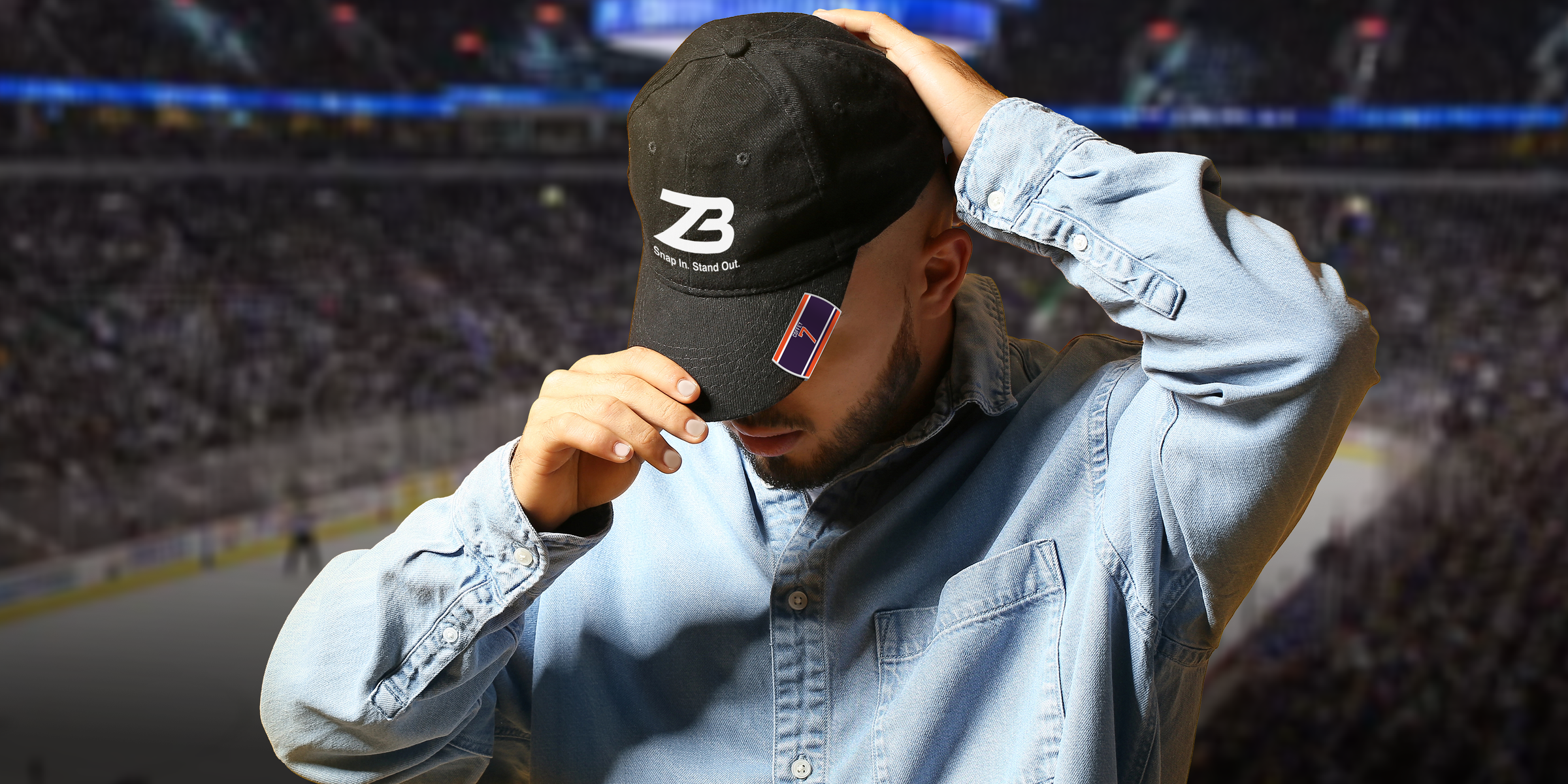 A person in a light denim shirt adjusts a black cap while standing in front of a blurred, crowded indoor stadium. The cap has a white ZB logo and a small sticker on the brim.