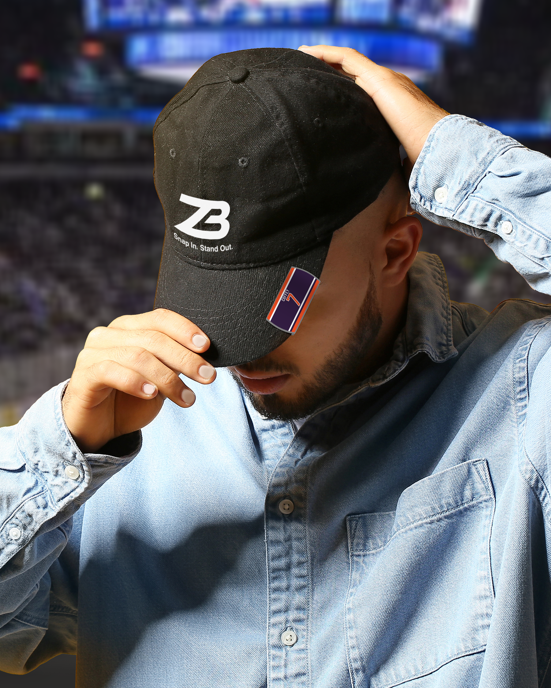 A person in a denim shirt tilts a black cap with the ZB logo and the words Stand Up. Stand Out. embroidered on it, in front of a blurred stadium background.