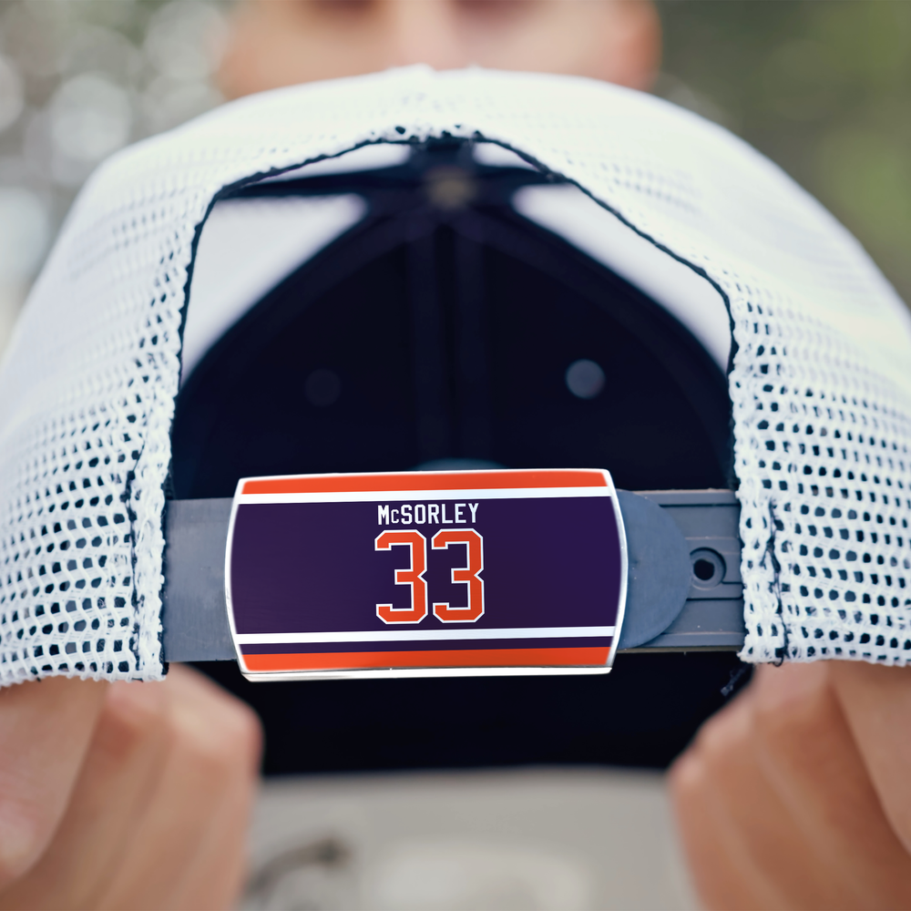 A person holds a white mesh ZippyBack™ baseball cap, showing the adjustable strap with a label reading Marty McSorley in white and orange text on a purple background.