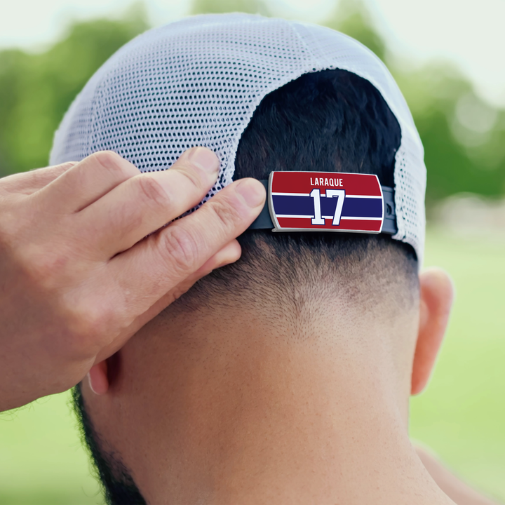 A person adjusts the strap of a white mesh ZippyBack™ cap, which features a red, white, and blue Georges Laraque nameplate with number 17—reflecting heavyweight impact. Blurred green trees fill the background.