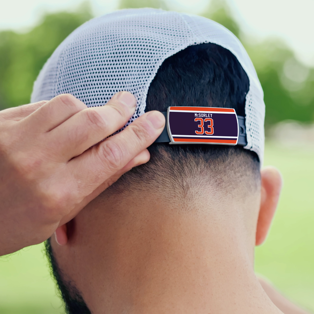 Someone adjusts a white ZippyBack™ cap called "Marty McSorley," featuring a nameplate with “MSRDRLEY 33” on the back strap. Green trees and blurred outdoor scenery appear in the background.