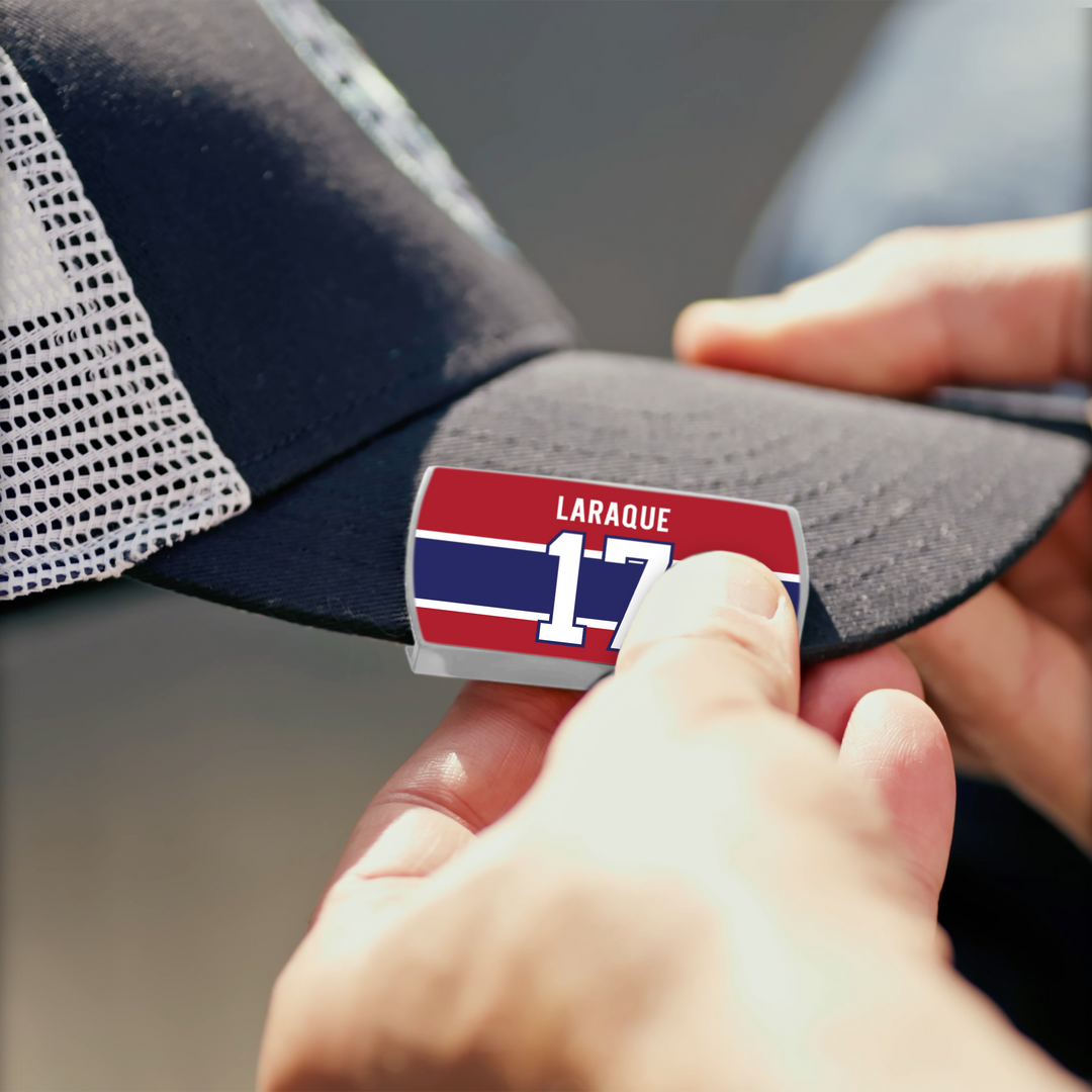A person adds a ZippyBack™ Georges Laraque red, blue, and white pin—featuring "Laraque" and number 17—to a black mesh baseball cap, symbolizing heavyweight strength and discipline.