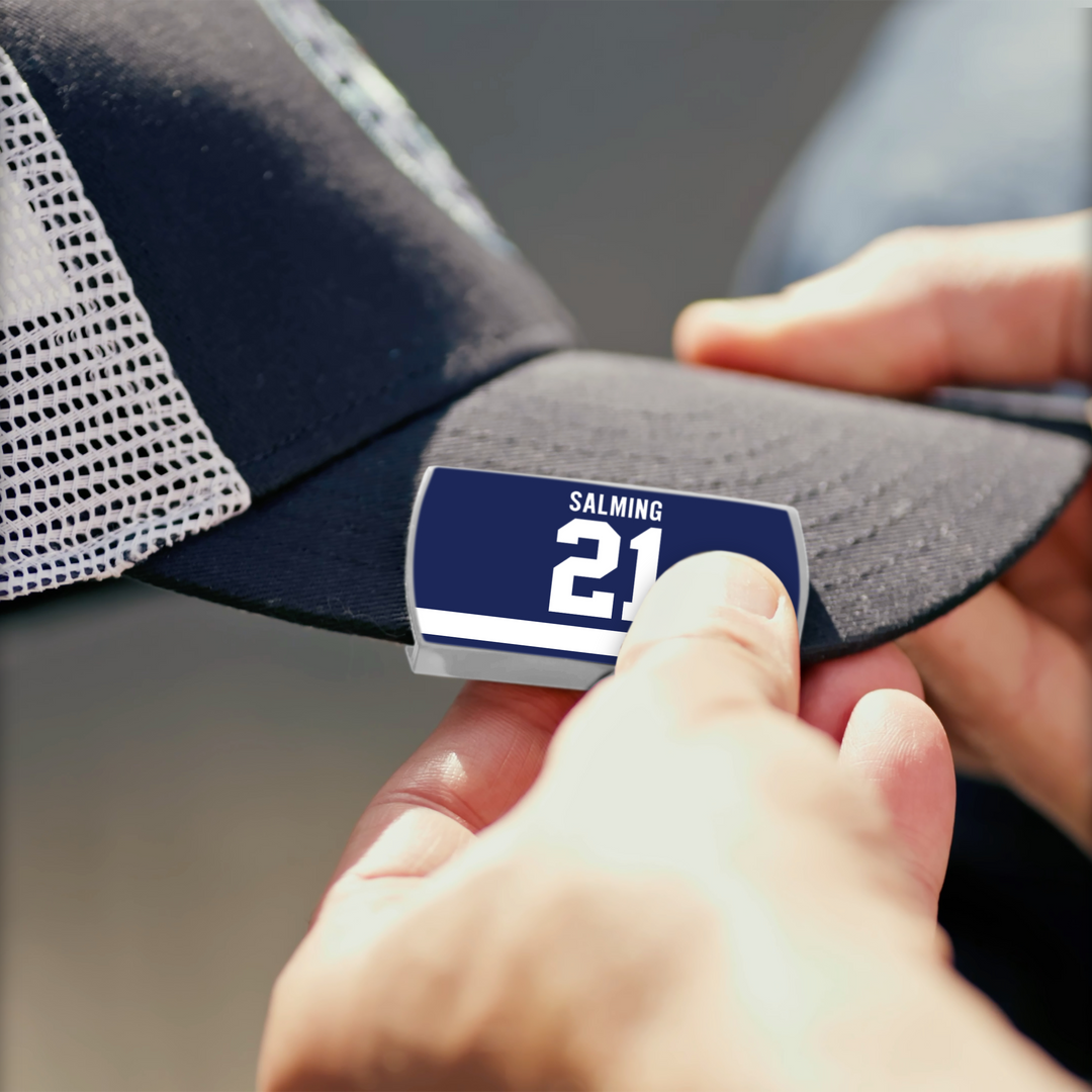 Close-up of hands attaching a rectangular blue label with white text and two stripes—featuring Börje Salming—onto a dark mesh ZippyBack™ baseball cap.