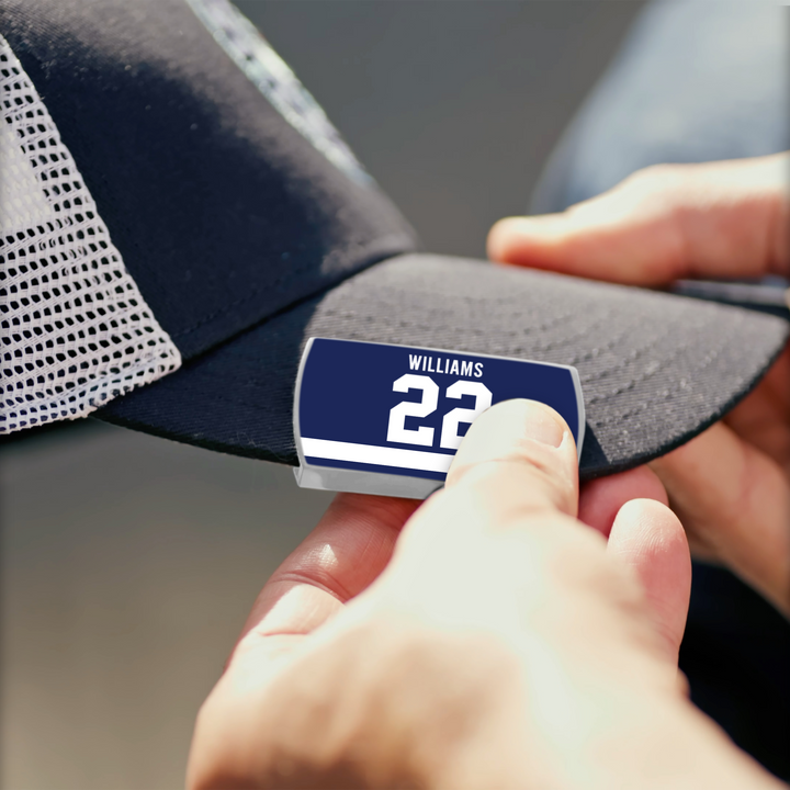 A person attaches a blue and white WILLIAMS 22 label to the brim of a black and white mesh baseball cap from ZippyBack™, channeling Tiger Williams’ fearless play.