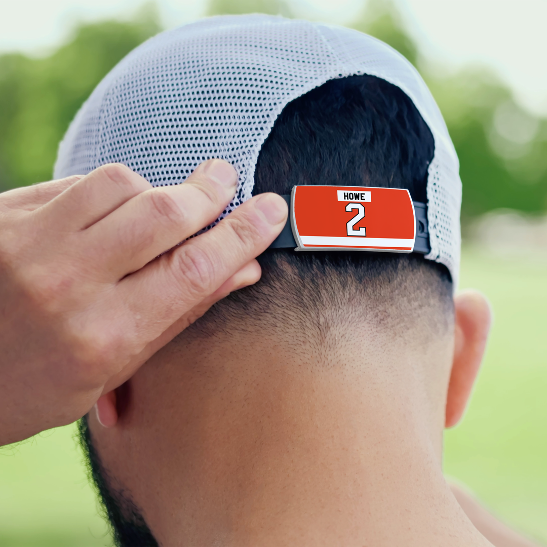 A person adjusts a ZippyBack™ Mark Howe white mesh baseball cap worn backwards, featuring a red clip on the back strap with "HOME" and the number 2, in front of blurred greenery.