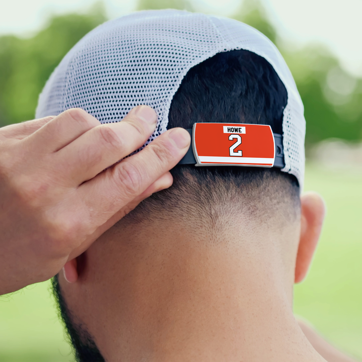 A person adjusts a ZippyBack™ Mark Howe white mesh baseball cap worn backwards, featuring a red clip on the back strap with "HOME" and the number 2, in front of blurred greenery.
