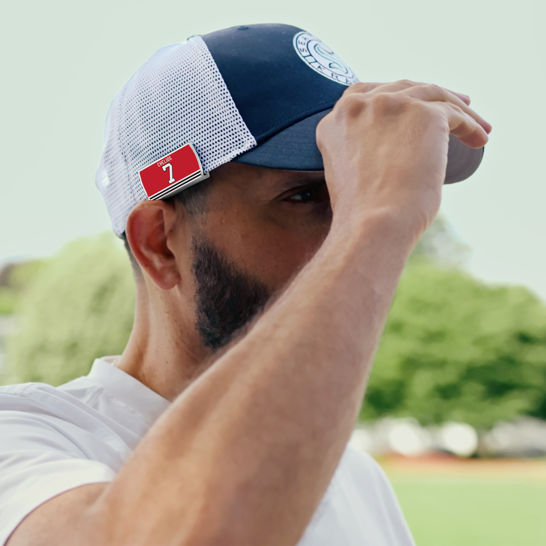 A bearded man in a navy and white ZippyBack™ Chris Chelios mesh cap holds the brim. A red tag with the number 7 nods to hockey defenseman durability; blurred trees and greenery are in the background.