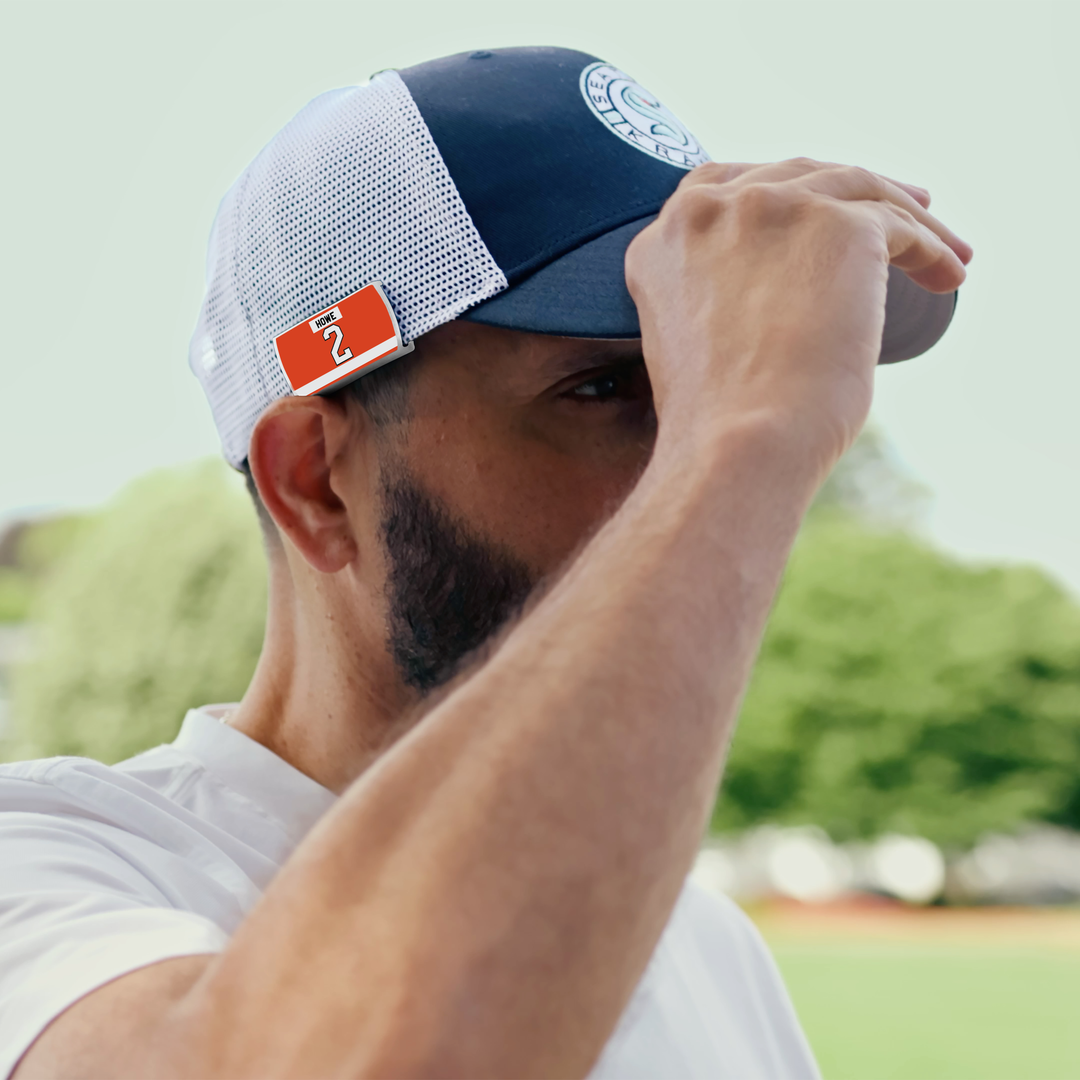 A man adjusts the brim of a ZippyBack™ Mark Howe navy and white mesh cap with a red tag while standing outdoors, with blurred green trees in the background.