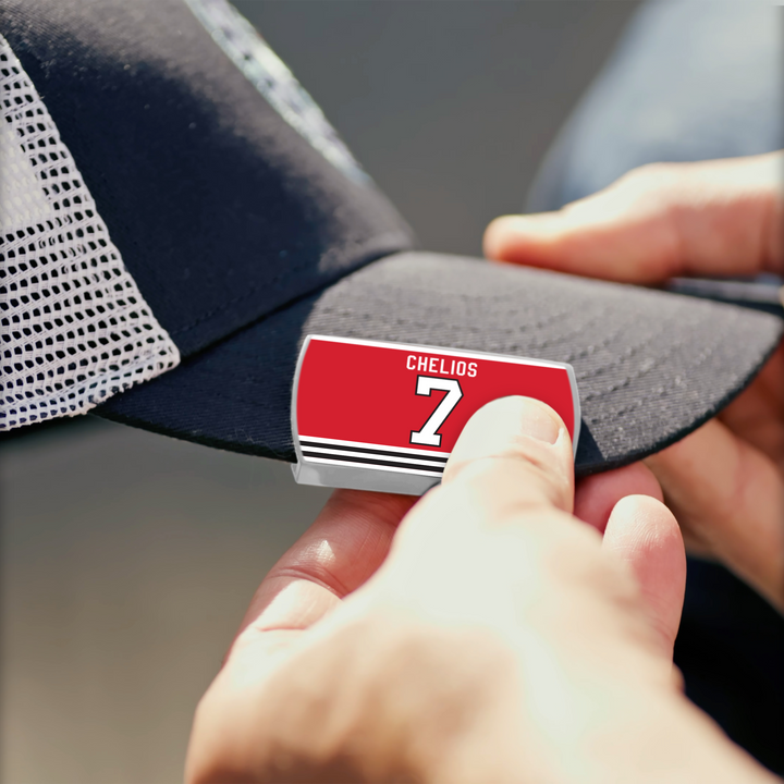Someone attaches a ZippyBack™ Chris Chelios pin, red and white with his name and 7 in bold white text, to a black mesh baseball cap—honoring his legendary durability and physical play on the ice.