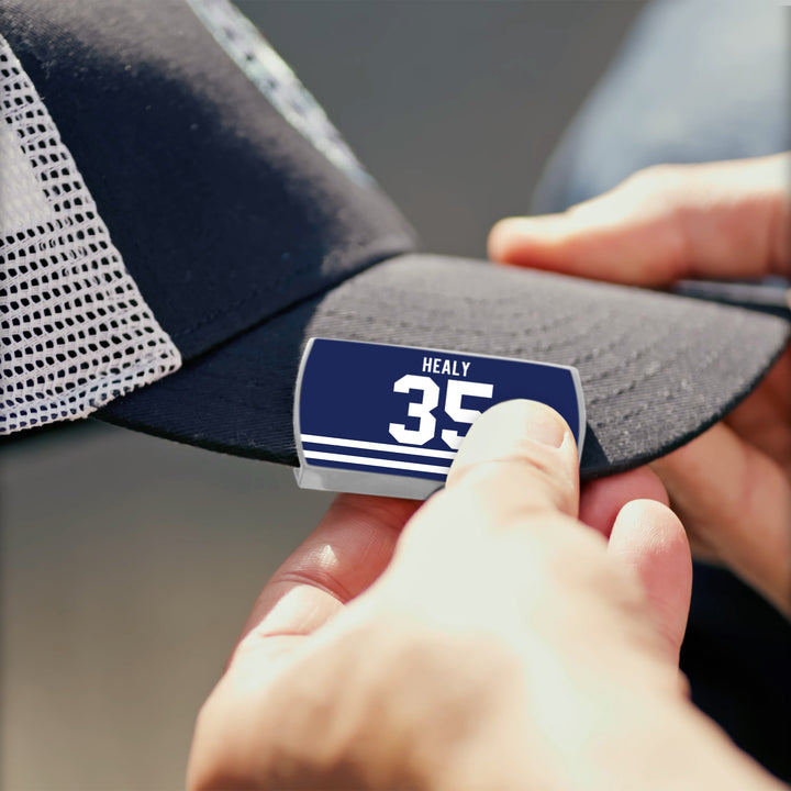 Close-up of hands attaching a blue Glenn Healy name tag, honoring the NHL goaltender, to the brim of a black and white mesh ZippyBack™ baseball cap.
