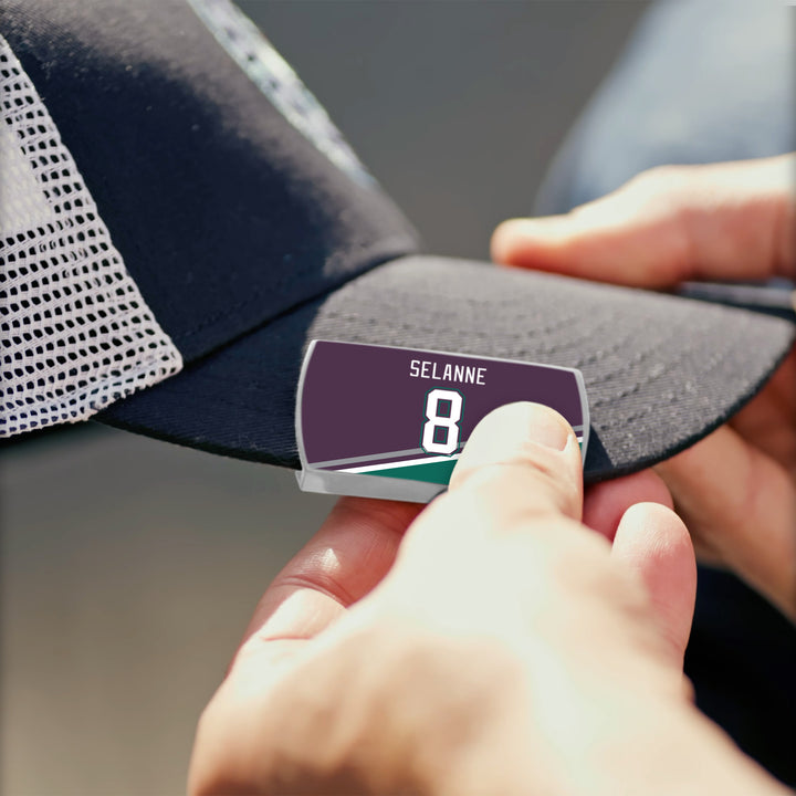 A person attaches a ZippyBack™ nameplate featuring Teemu Selänne and number 8 to the brim of a black and white mesh baseball cap.