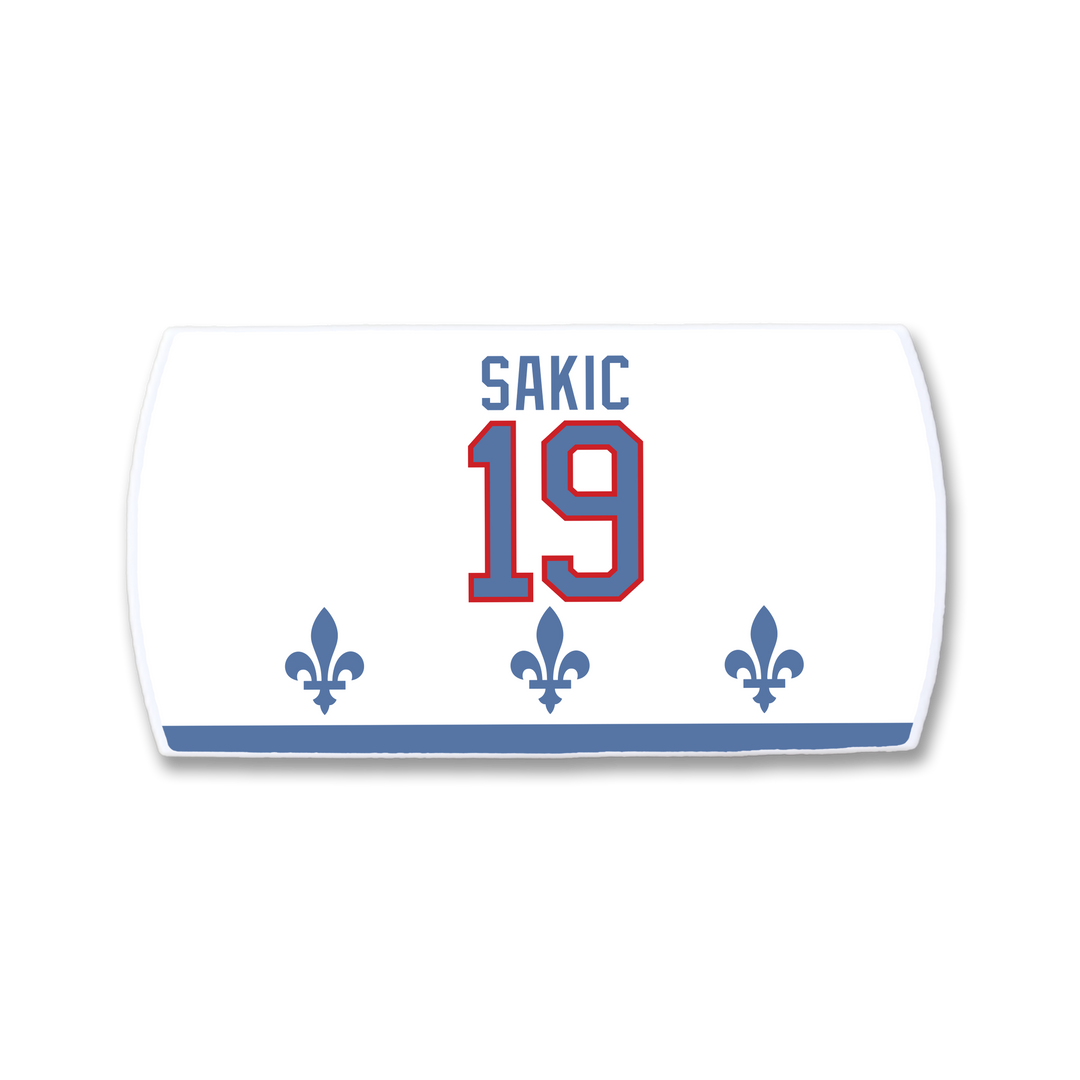 The ZippyBack™ Joe Sakic white hockey jersey nameplate features “SAKIC” in blue above red number 19, three blue fleur-de-lis below, and a blue stripe at the bottom—honoring the legendary player.
