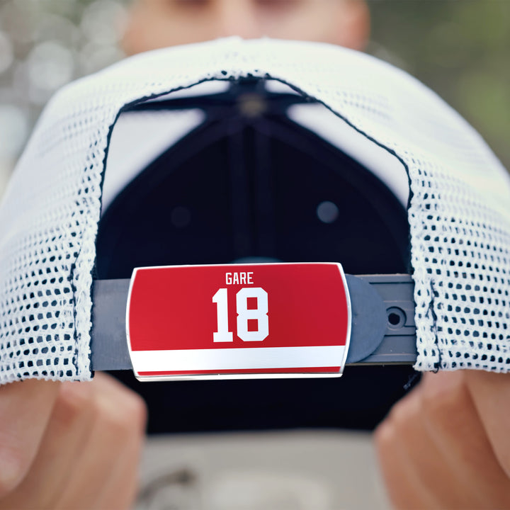 A person holds a white mesh ZippyBack™ baseball cap backwards, displaying a red “Danny Gare 18” label on the strap; their face is blurred in the background.