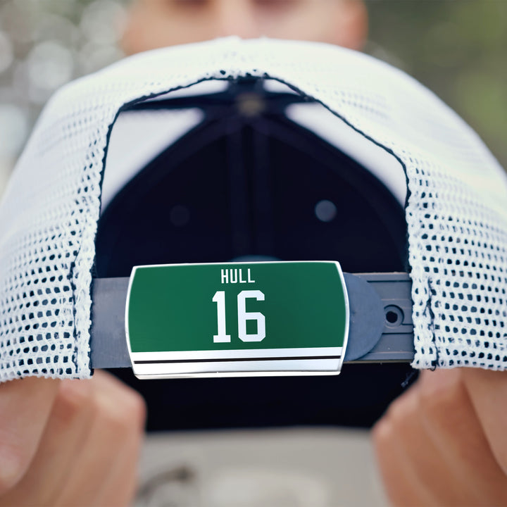 A person holds a ZippyBack™ Brett Hull cap—a white mesh baseball cap featuring a green nameplate with HULL 16 in white. The background is blurred, highlighting the product’s tribute to the famed goal scorer.