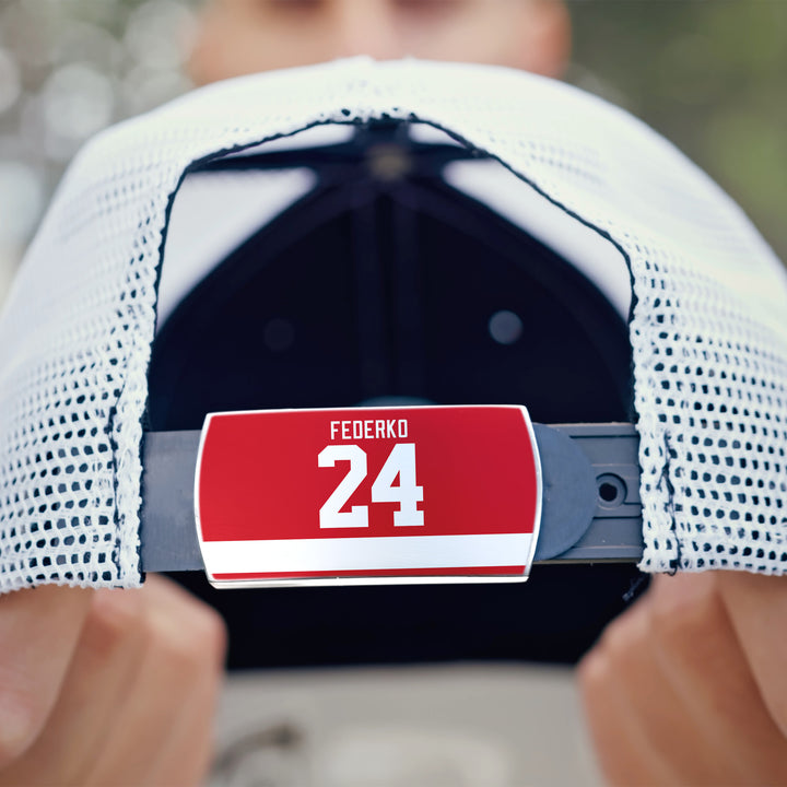 A close-up of a person holding the adjustable strap of a white mesh ZippyBack™ baseball cap named "Bernie Federko," featuring a red label with "FEQERKO" and the number 24 in white—evoking quiet control on the ice.