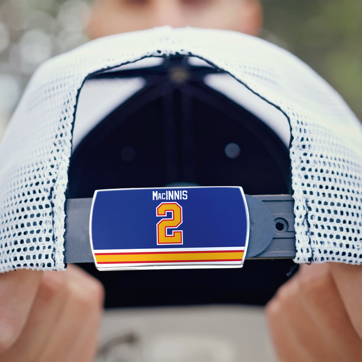 A close-up of the ZippyBack™ Al MacInnis white mesh cap shows a blue nameplate with “MacINNIS” and the number 2 in yellow and red on the adjustable strap, held by a person’s hands.