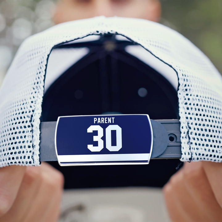 A close-up of a person holding a white ZippyBack™ mesh baseball cap named "Bernie Parent," highlighting its adjustable strap with a blue label reading PARENT 30. The face is out of focus in the background.