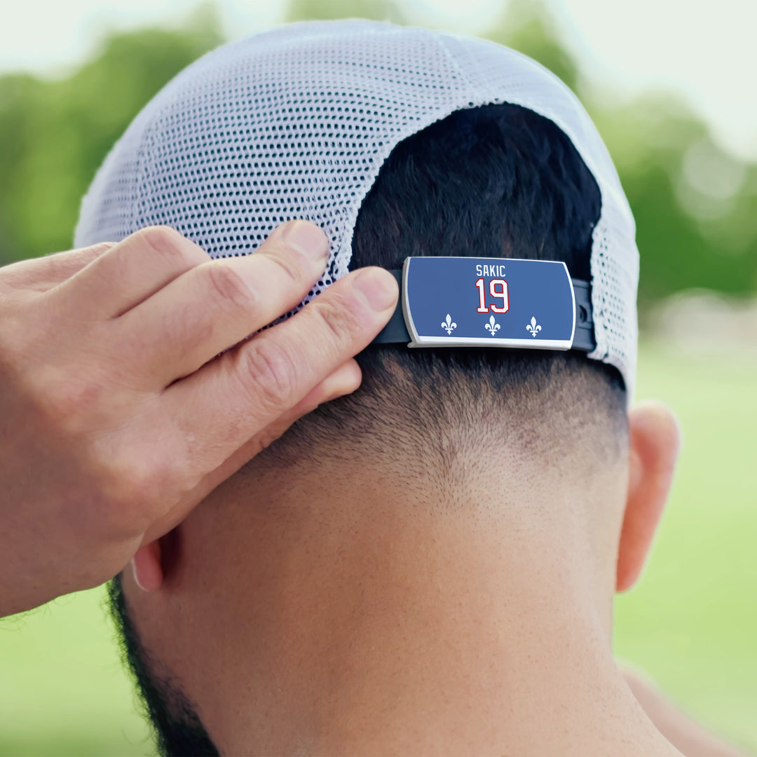 Outdoors, a person adjusts the back of a ZippyBack™ Joe Sakic mesh baseball cap, featuring his name, number 19, and fleur-de-lis symbols on the clip, with blurred green trees behind.