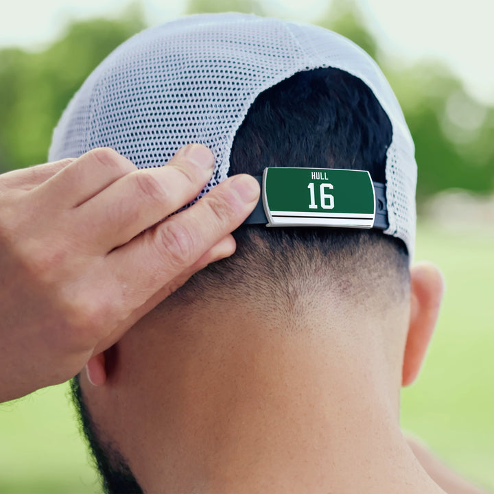 A person adjusts a white mesh baseball cap with a ZippyBack™ digital device clipped to the back, displaying "Brett Hull" on a green screen. The blurred outdoor background nods to Hull’s legendary precision.