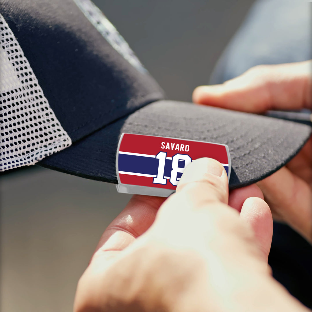 A person clips a ZippyBack™ Denis Savard red, white, and blue badge to the brim of a black mesh baseball cap.
