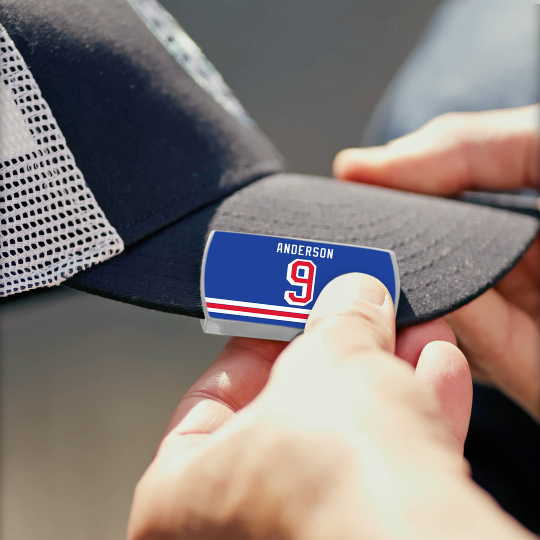A close-up of hands attaching the Glenn Anderson name tag by ZippyBack™—featuring a blue label, red number 9, and white stripes—onto a black and white mesh baseball cap.