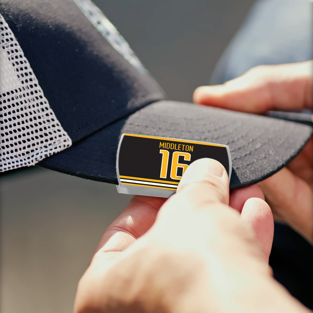 A person puts a rectangular "Rick Middleton" label in black, yellow, and white on the brim of a black-and-white ZippyBack™ trucker hat, celebrating the offensive legend.