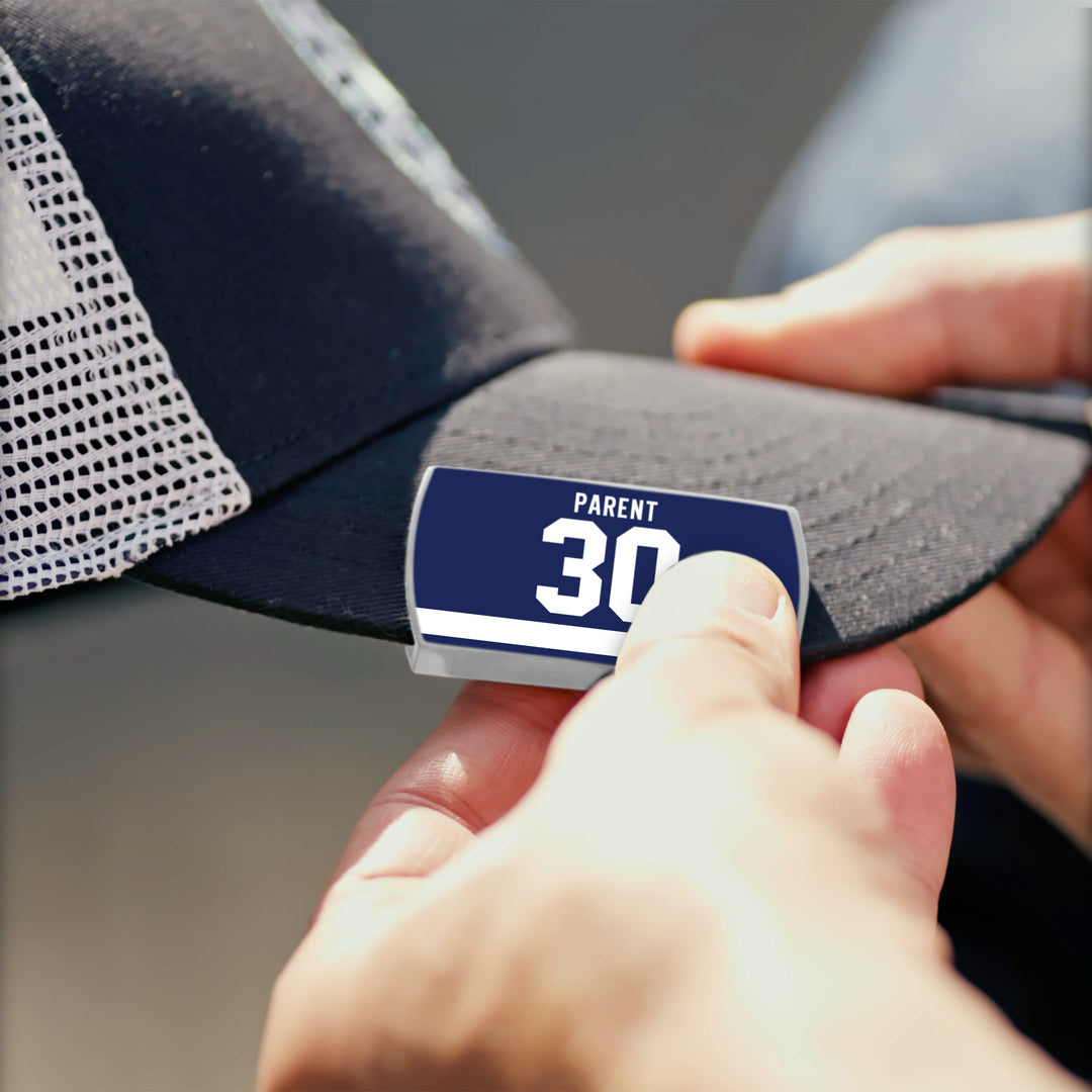 A person attaches a ZippyBack™ Bernie Parent blue and white name tag labeled PARENT 30 to the brim of a black mesh baseball cap, celebrating Flyers’ championship-era goaltending.
