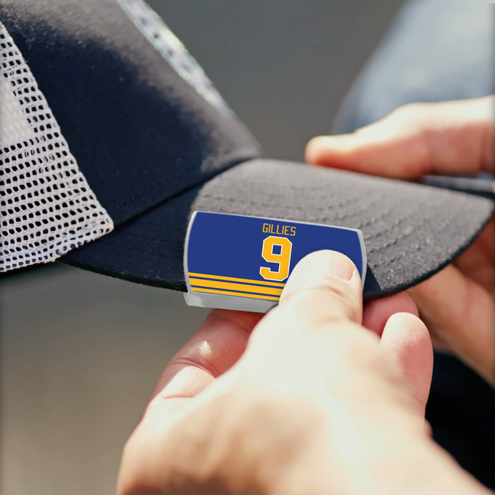 A person attaches a blue and yellow “Clark Gillies” ZippyBack™ pin to the brim of a black mesh baseball cap, honoring the legendary power forward.