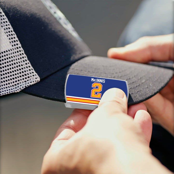 A person attaches the ZippyBack™ Al MacInnis rectangular pin, featuring 2 and blue, orange, white, and yellow stripes at the bottom, to a black mesh baseball cap to honor the hockey legend known for his powerful slapshot.