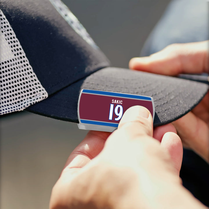 A person attaches a ZippyBack™ Joe Sakic label to the brim of a black and white mesh baseball cap.