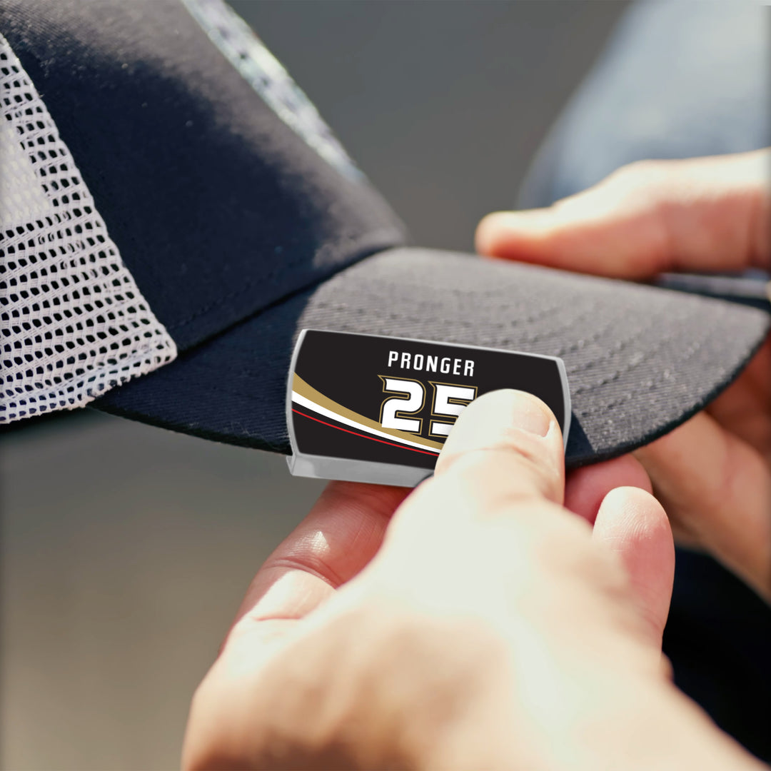 A person applies a ZippyBack™ Chris Pronger sticker—featuring “PRONGER 25” and black, gold, and white stripes—onto a black mesh baseball cap to honor the legendary defenseman’s puck-moving skills.