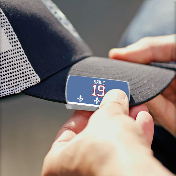 A person attaches a blue rectangular Joe Sakic patch with SAKIC 19 and two fleur-de-lis symbols to the brim of a black and white ZippyBack™ mesh baseball cap.