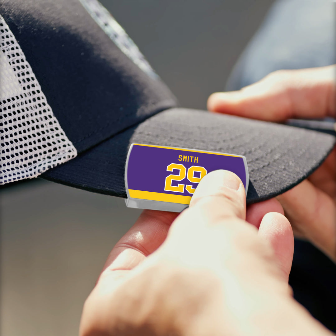 A person attaches a ZippyBack™ name tag with "Billy Smith 29" in yellow on purple to the brim of a black and white mesh baseball cap, celebrating Billy Smith’s clutch hockey performances.