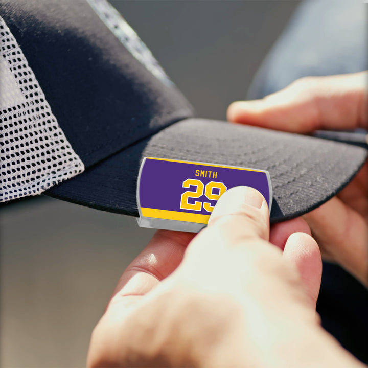 A person attaches a ZippyBack™ name tag with "Billy Smith 29" in yellow on purple to the brim of a black and white mesh baseball cap, celebrating Billy Smith’s clutch hockey performances.