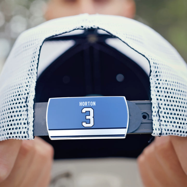 A hand displays a white mesh ZippyBack™ Tim Horton baseball cap, highlighting the adjustable strap with a blue HORTON 3 label. The background is blurred.