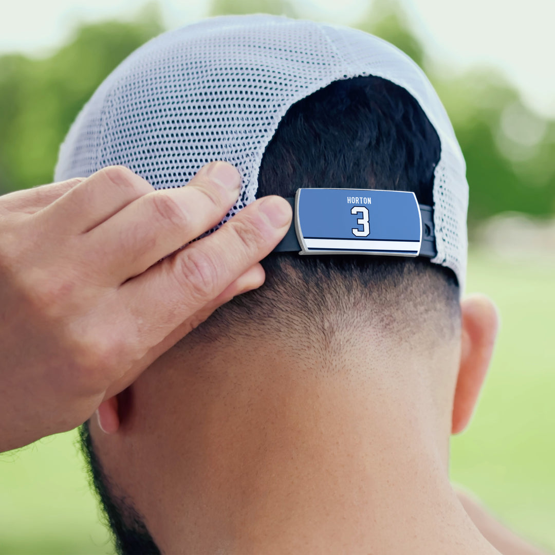 A person adjusts the strap of a white mesh ZippyBack™ cap featuring a blue "Tim Horton" nameplate and the number 3, with green trees blurred in the background.
