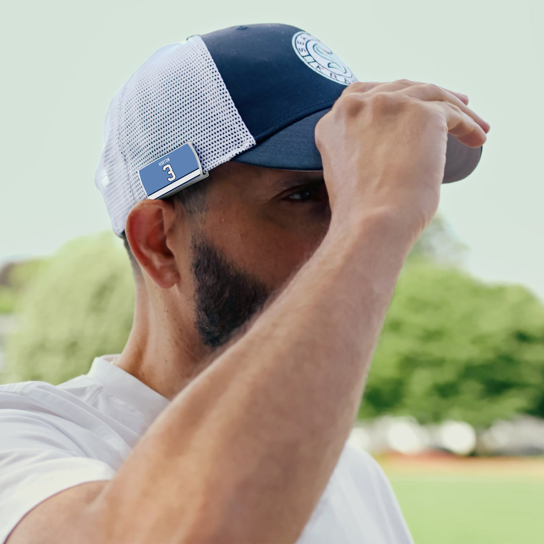 A bearded man wears a ZippyBack™ Tim Horton mesh baseball cap with a number 3 patch and a white shirt, shielding his eyes outdoors among green trees and a blurred background.