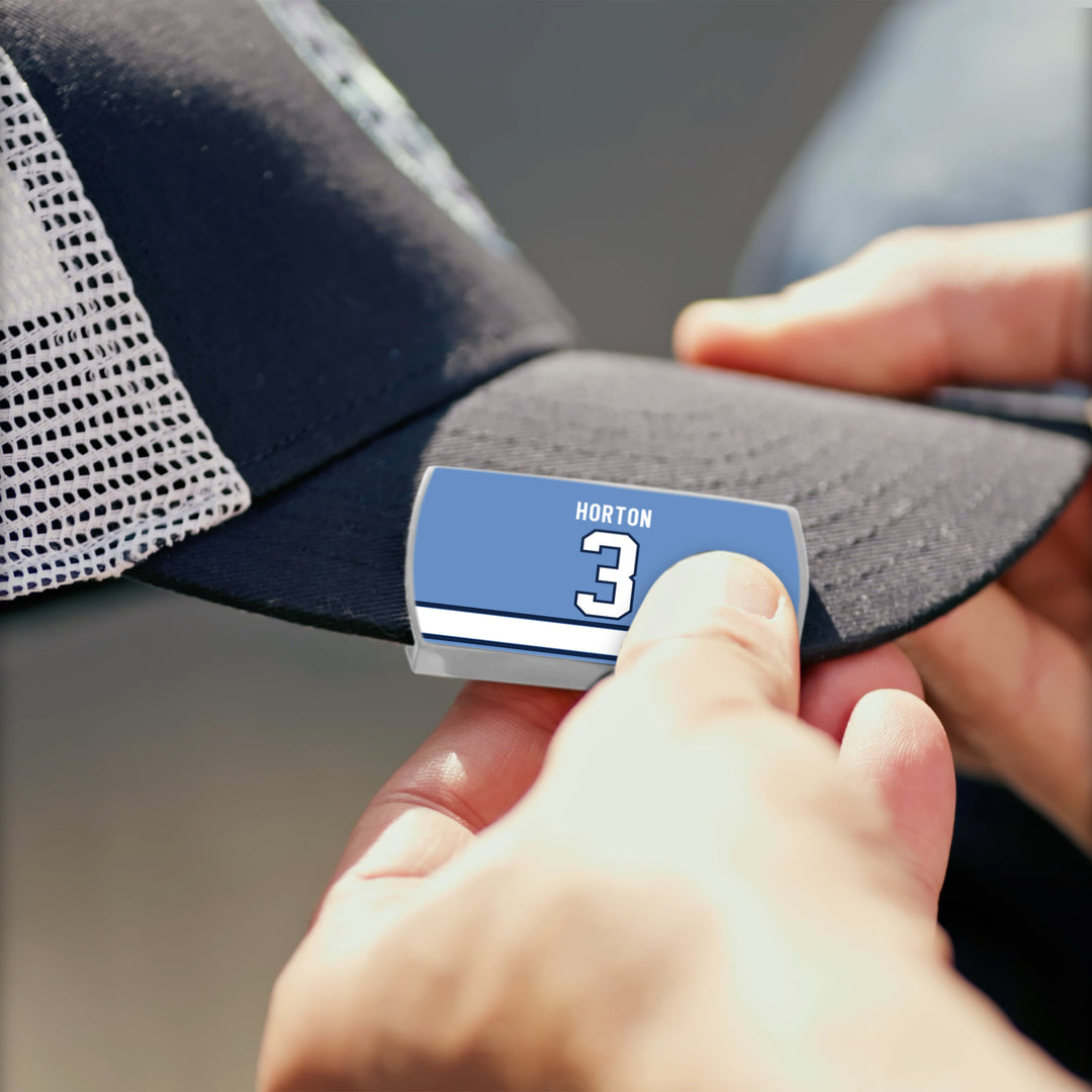 A person attaches a blue and white ZippyBack™ label reading Tim Horton to the brim of a dark mesh baseball cap, honoring legendary hockey defenseman Tim Horton.