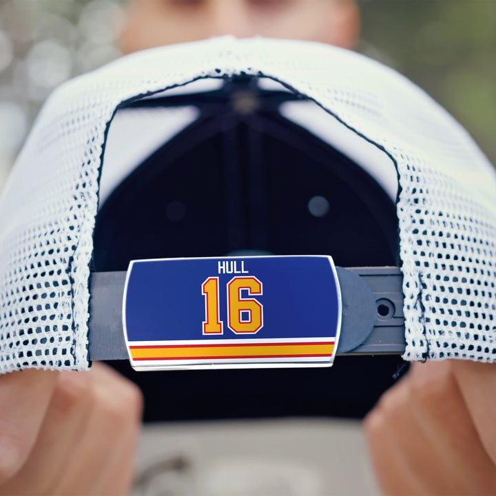 A close-up shows a ZippyBack™ Brett Hull mesh baseball cap’s adjustable strap with a blue “HULL 16” sticker in yellow and red stripes, honoring Brett Hull, as someone holds the cap.
